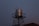 A metallic water tank is elevated on a black lattice stand against a dimly lit evening sky. The tank reflects a subtle amount of light, creating a contrast with the darker surroundings. The roof of a building is partially visible at the base with a weather vane or antenna on top.