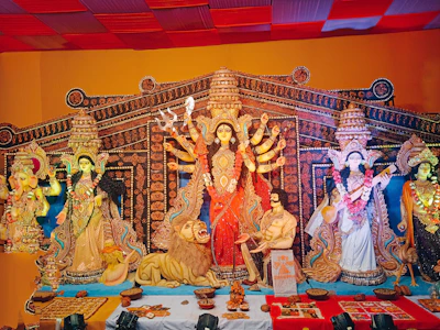 A vibrant and intricately decorated tableau of Hindu deities arranged in a traditional setting. The central figure is a multi-armed goddess adorned with a red sari, surrounded by other deities and a lion. There are offerings and decorative elements placed in front of the figures.