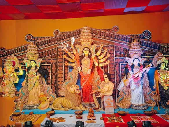 A vibrant and intricately decorated tableau of Hindu deities arranged in a traditional setting. The central figure is a multi-armed goddess adorned with a red sari, surrounded by other deities and a lion. There are offerings and decorative elements placed in front of the figures.