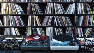 A collection of vinyl records is neatly organized on shelves in the background, while DJ equipment including two turntables and a mixer is positioned in the foreground. The turntables are equipped with red vinyl records.