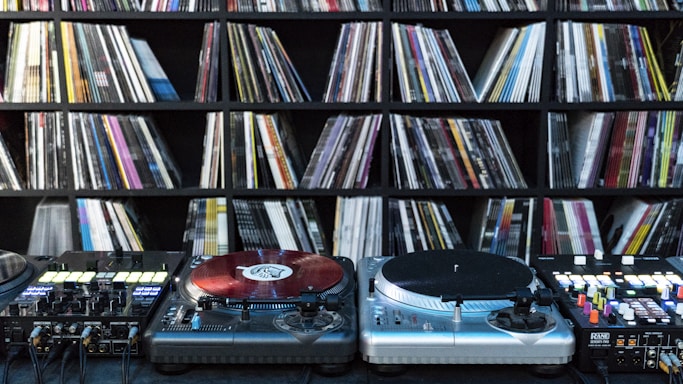 A collection of vinyl records is neatly organized on shelves in the background, while DJ equipment including two turntables and a mixer is positioned in the foreground. The turntables are equipped with red vinyl records.