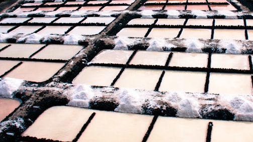 Multiple rectangular salt pans arranged in a grid pattern with dark embankments. The surfaces of the pans have varying shades of brown and beige with salt deposits accumulated at the edges.