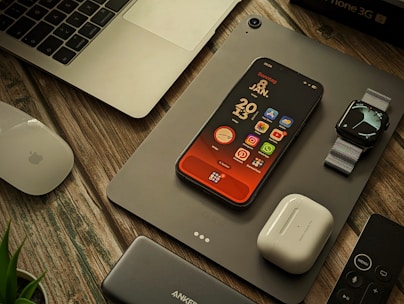 A collection of Apple products and accessories are arranged on a wooden surface. There is a MacBook partially visible, an iPhone with a colorful home screen, an Apple Watch with a metallic band, a pair of AirPods, a Magic Mouse, a power bank, and a remote control. A plant is slightly visible in the bottom left corner.
