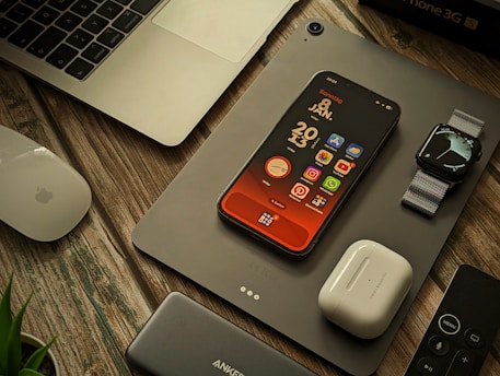A collection of Apple products and accessories are arranged on a wooden surface. There is a MacBook partially visible, an iPhone with a colorful home screen, an Apple Watch with a metallic band, a pair of AirPods, a Magic Mouse, a power bank, and a remote control. A plant is slightly visible in the bottom left corner.