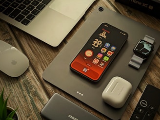 A collection of Apple products and accessories are arranged on a wooden surface. There is a MacBook partially visible, an iPhone with a colorful home screen, an Apple Watch with a metallic band, a pair of AirPods, a Magic Mouse, a power bank, and a remote control. A plant is slightly visible in the bottom left corner.