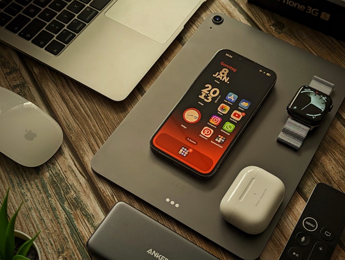 A collection of Apple products and accessories are arranged on a wooden surface. There is a MacBook partially visible, an iPhone with a colorful home screen, an Apple Watch with a metallic band, a pair of AirPods, a Magic Mouse, a power bank, and a remote control. A plant is slightly visible in the bottom left corner.