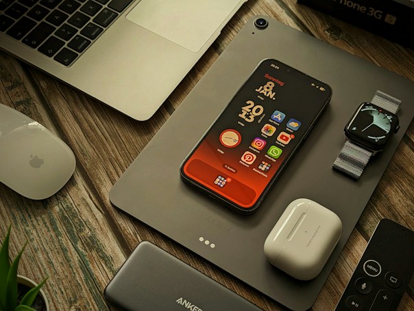 A collection of Apple products and accessories are arranged on a wooden surface. There is a MacBook partially visible, an iPhone with a colorful home screen, an Apple Watch with a metallic band, a pair of AirPods, a Magic Mouse, a power bank, and a remote control. A plant is slightly visible in the bottom left corner.