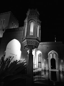 An ornate, traditional-style lamp is prominently featured in the foreground, surrounded by lush foliage. The background consists of an architectural structure with arched doorways, illuminated softly against the night sky, creating a serene and timeless ambiance.