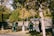 Several people wearing green uniforms and orange hats are cleaning a park area. They appear to be using brooms and bags to gather fallen leaves. The park is lush with trees and greenery, and the sunlight casts dappled shadows on the ground.