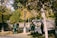 Several people wearing green uniforms and orange hats are cleaning a park area. They appear to be using brooms and bags to gather fallen leaves. The park is lush with trees and greenery, and the sunlight casts dappled shadows on the ground.