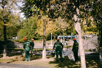 Several people wearing green uniforms and orange hats are cleaning a park area. They appear to be using brooms and bags to gather fallen leaves. The park is lush with trees and greenery, and the sunlight casts dappled shadows on the ground.