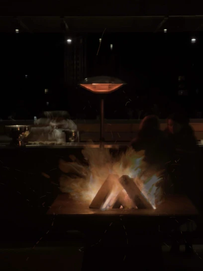 A nighttime scene featuring an outdoor setting with a fire pit at the center. Logs are arranged in the fire pit with flames and sparks emanating from them, providing warm illumination. In the background, there are blurred figures seated, along with an outdoor heater, suggesting a cozy gathering.