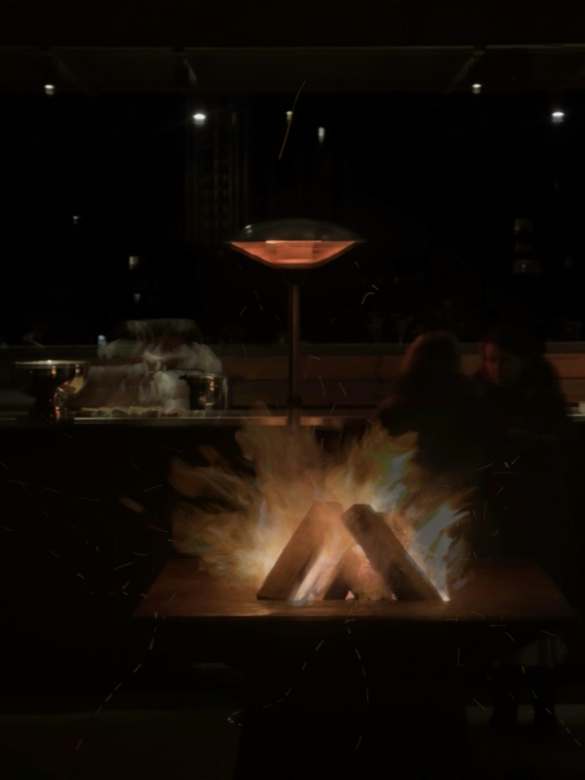 A cozy backyard evening scene showing the large brozero brasero glowing warmly surrounded by friends enjoying grilled meat.