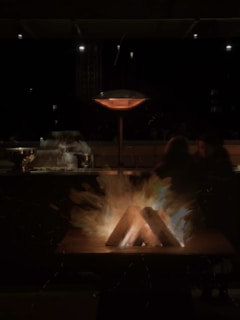 A nighttime scene featuring an outdoor setting with a fire pit at the center. Logs are arranged in the fire pit with flames and sparks emanating from them, providing warm illumination. In the background, there are blurred figures seated, along with an outdoor heater, suggesting a cozy gathering.