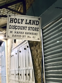 A sign for the Holy Land Discount Store, displaying the proprietor's name M. Nafez Shweiki, and address at David Street No. 61. The sign is hung in a dimly lit alleyway with closed metal shutters and weathered wall structures.
