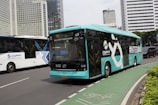A turquoise electric bus, part of the TransJakarta fleet, is traveling on a city road alongside other vehicles. It features modern design elements and branding on its side. The road is lined with tall modern office buildings in the background, and there is a designated bike lane visible in the foreground.