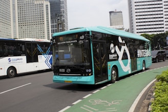 A turquoise electric bus, part of the TransJakarta fleet, is traveling on a city road alongside other vehicles. It features modern design elements and branding on its side. The road is lined with tall modern office buildings in the background, and there is a designated bike lane visible in the foreground.
