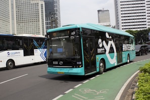 A turquoise electric bus, part of the TransJakarta fleet, is traveling on a city road alongside other vehicles. It features modern design elements and branding on its side. The road is lined with tall modern office buildings in the background, and there is a designated bike lane visible in the foreground.