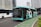A turquoise electric bus, part of the TransJakarta fleet, is traveling on a city road alongside other vehicles. It features modern design elements and branding on its side. The road is lined with tall modern office buildings in the background, and there is a designated bike lane visible in the foreground.
