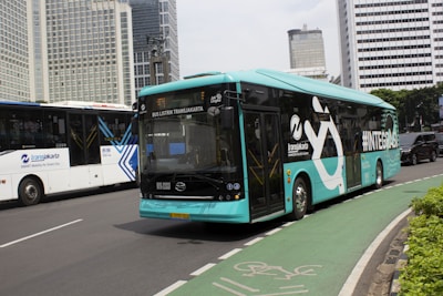 A turquoise electric bus, part of the TransJakarta fleet, is traveling on a city road alongside other vehicles. It features modern design elements and branding on its side. The road is lined with tall modern office buildings in the background, and there is a designated bike lane visible in the foreground.