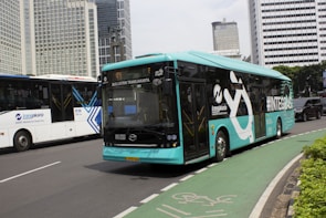 A turquoise electric bus, part of the TransJakarta fleet, is traveling on a city road alongside other vehicles. It features modern design elements and branding on its side. The road is lined with tall modern office buildings in the background, and there is a designated bike lane visible in the foreground.
