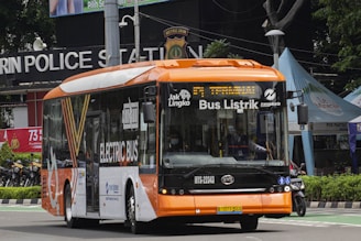 A modern electric bus with a bright orange and white color scheme is driving along a street. The bus displays 'Bus Listrik' and 'Electric Bus' on its side and front, indicating it is powered by electricity. In the background, a police station sign and various tents or kiosks can be seen.