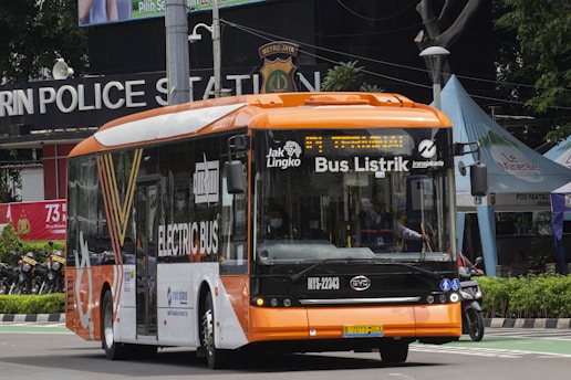 A modern electric bus with a bright orange and white color scheme is driving along a street. The bus displays 'Bus Listrik' and 'Electric Bus' on its side and front, indicating it is powered by electricity. In the background, a police station sign and various tents or kiosks can be seen.