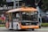 A modern electric bus with a bright orange and white color scheme is driving along a street. The bus displays 'Bus Listrik' and 'Electric Bus' on its side and front, indicating it is powered by electricity. In the background, a police station sign and various tents or kiosks can be seen.