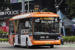 A modern electric bus with a bright orange and white color scheme is driving along a street. The bus displays 'Bus Listrik' and 'Electric Bus' on its side and front, indicating it is powered by electricity. In the background, a police station sign and various tents or kiosks can be seen.