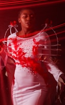 A person wearing a white dress with decorative red feathers and pearl-like accents, surrounded by flowing red fabric that creates a dynamic and dreamy effect.