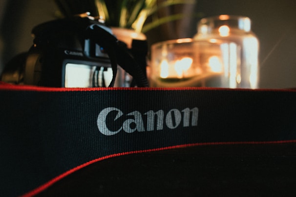 A Canon camera is partially visible with a focus on a Canon-branded strap in the foreground. In the background, a blurred scene includes lit candles inside glass containers, creating a warm, atmospheric glow. Some green plant leaves can also be seen.