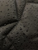 Close-up view of a dark fabric, possibly a jacket, with water droplets beading up on the surface. The fabric has prominent stitching lines forming a geometric pattern.