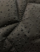 Close-up view of a dark fabric, possibly a jacket, with water droplets beading up on the surface. The fabric has prominent stitching lines forming a geometric pattern.
