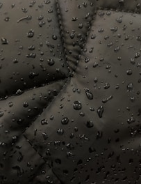 Close-up view of a dark fabric, possibly a jacket, with water droplets beading up on the surface. The fabric has prominent stitching lines forming a geometric pattern.