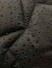 Close-up view of a dark fabric, possibly a jacket, with water droplets beading up on the surface. The fabric has prominent stitching lines forming a geometric pattern.