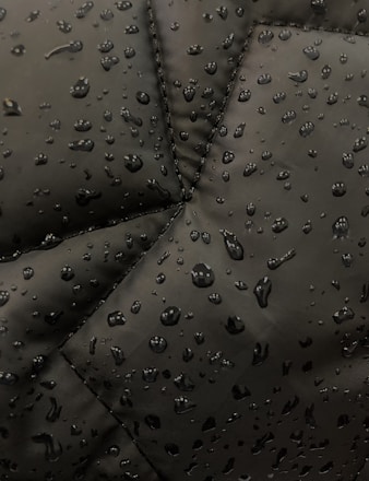 Close-up view of a dark fabric, possibly a jacket, with water droplets beading up on the surface. The fabric has prominent stitching lines forming a geometric pattern.