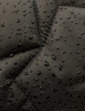 Close-up view of a dark fabric, possibly a jacket, with water droplets beading up on the surface. The fabric has prominent stitching lines forming a geometric pattern.