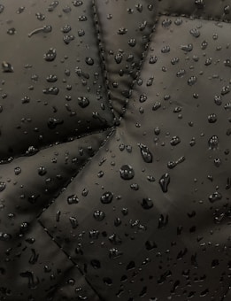 Close-up view of a dark fabric, possibly a jacket, with water droplets beading up on the surface. The fabric has prominent stitching lines forming a geometric pattern.
