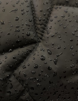 Close-up view of a dark fabric, possibly a jacket, with water droplets beading up on the surface. The fabric has prominent stitching lines forming a geometric pattern.