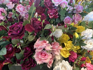 A bouquet of vibrant artificial roses featuring a variety of colors, including deep red, pink, white, and yellow. The roses are arranged closely together, with green leaves interspersed throughout, creating a lush and abundant floral display.