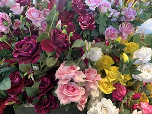 A bouquet of vibrant artificial roses featuring a variety of colors, including deep red, pink, white, and yellow. The roses are arranged closely together, with green leaves interspersed throughout, creating a lush and abundant floral display.