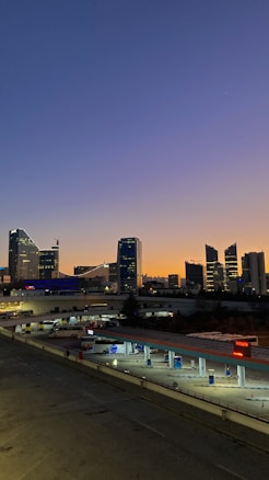 A cityscape photograph capturing a sunset behind a modern skyline with tall buildings silhouetted against the gradient sky. Below, a gas station with a few vehicles parked provides foreground interest, while lights begin to illuminate as daylight fades.