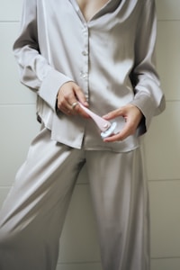 A person in a silky, long-sleeved pajama set is holding a pink and white razor. The pajamas have a shiny texture, and the setting appears indoors with a neutral, tiled background.