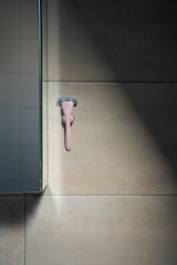 A pink razor is mounted on a beige tiled wall, with light casting shadows on the surface. Nearby, a section of a glass panel reflects part of the wall.
