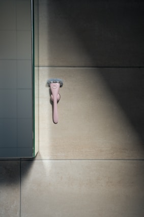A pink razor is mounted on a beige tiled wall, with light casting shadows on the surface. Nearby, a section of a glass panel reflects part of the wall.