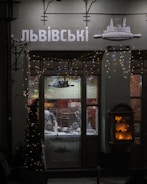 A dimly lit shop window displays a person inside, working on a counter with kitchen tools. Decorative fairy lights adorn the shopfront, and an illuminated display case is visible to the right. The signage above the window features Cyrillic lettering.