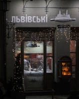 A dimly lit shop window displays a person inside, working on a counter with kitchen tools. Decorative fairy lights adorn the shopfront, and an illuminated display case is visible to the right. The signage above the window features Cyrillic lettering.
