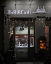 A dimly lit shop window displays a person inside, working on a counter with kitchen tools. Decorative fairy lights adorn the shopfront, and an illuminated display case is visible to the right. The signage above the window features Cyrillic lettering.