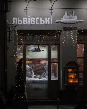 A dimly lit shop window displays a person inside, working on a counter with kitchen tools. Decorative fairy lights adorn the shopfront, and an illuminated display case is visible to the right. The signage above the window features Cyrillic lettering.