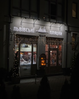 A modest storefront is softly illuminated in the evening with gentle string lights decorating the upper facade. There are display windows showcasing bakery goods inside, and through the glass, a person is seen working amidst trays and kitchenware. The street is dimly lit, and potted trees with small lights add a festive charm to the scene.